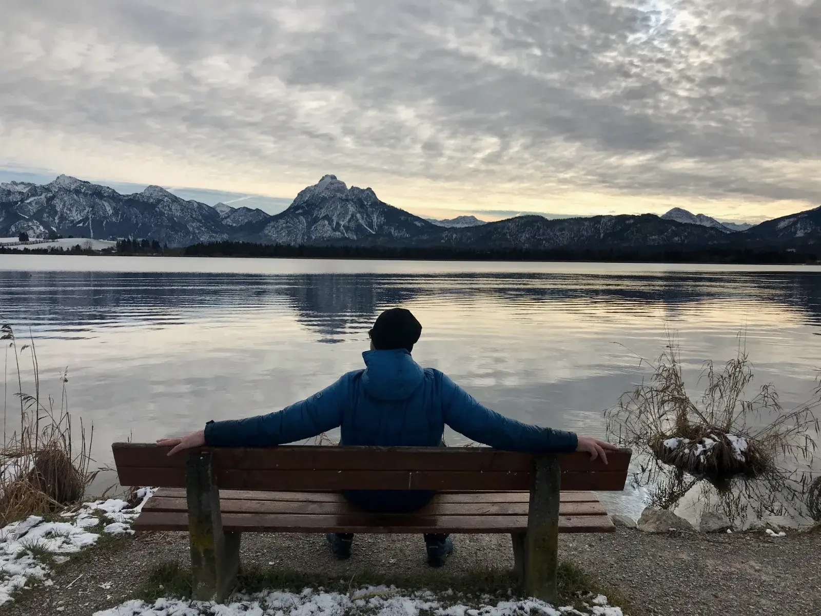 Andreas Hinz sitzt mit ausgebreiteten Armen auf einer Bank am Hopfensee und blickt auf die verschneiten Allgäuer Berge.